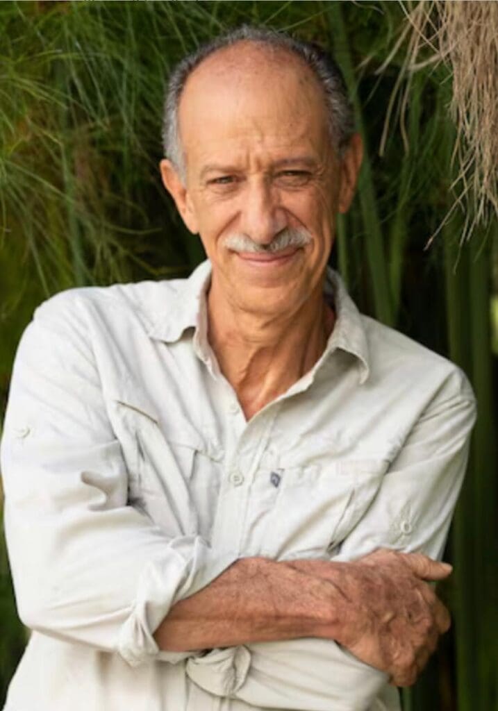 Roque Sevilla smiles with his arms crossed, wearing a light-colored long-sleeved shirt, in front of green vegetation.